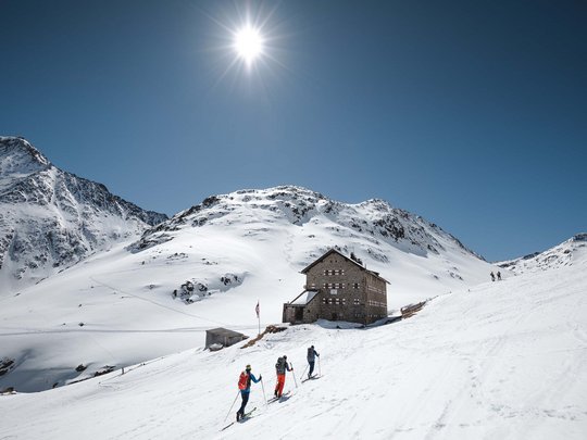 Skitouren rund um Vent: direkt von Ihrem Hotel in die Berge Drei Skifahrer vor einer Berghütte in verschneiter Alpenlandschaft bei Sonnenschein